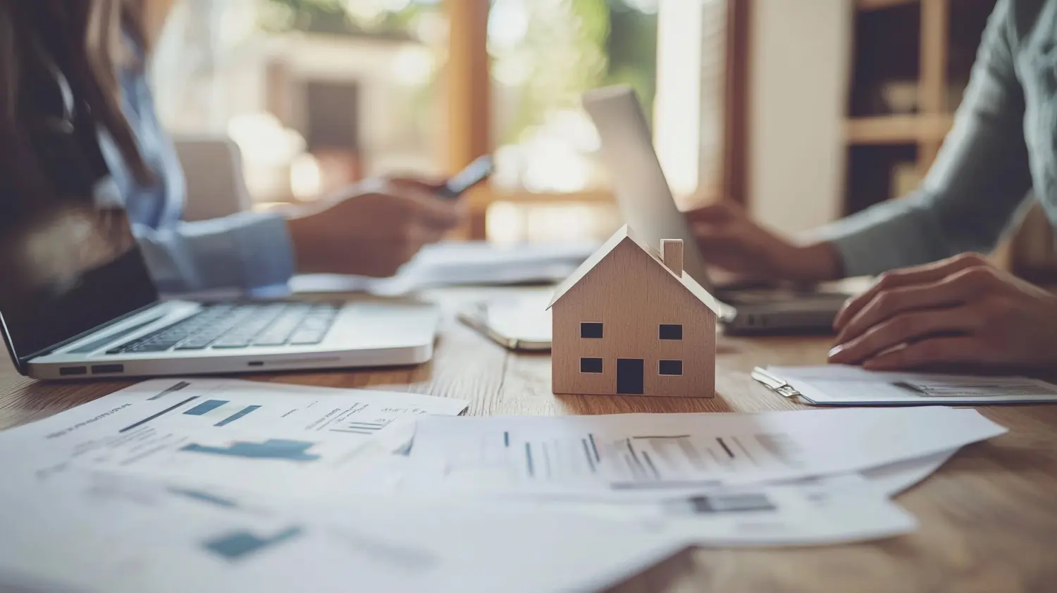 Ein gemeinsamer Arbeitsbereich mit einem Holzhausmodell auf Dokumenten, Laptops und Menschen, die über Immobilien oder Wohnthemen diskutieren.